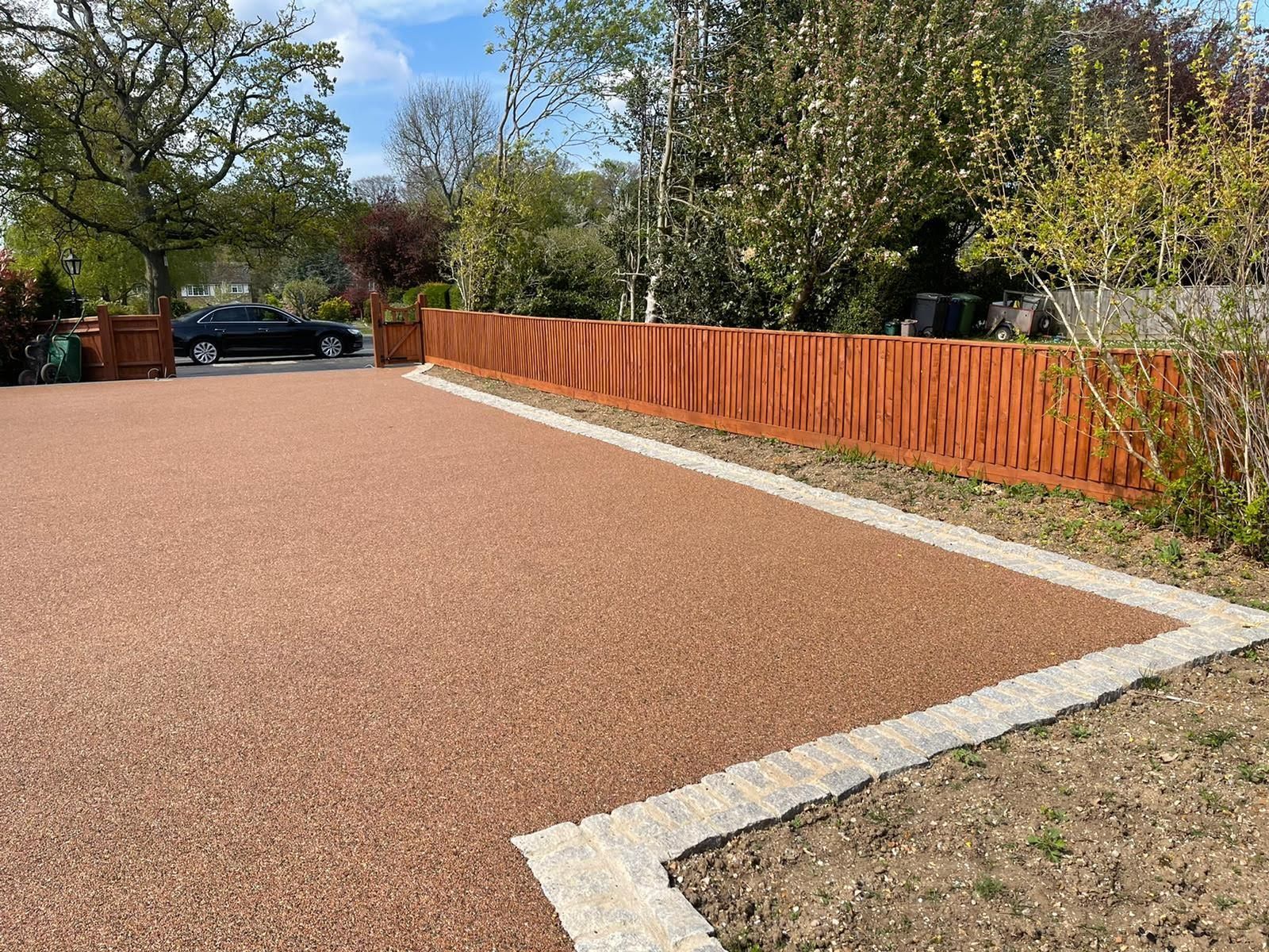 Red gravel driveway with orange wooden fence, bordered by light-colored stones.