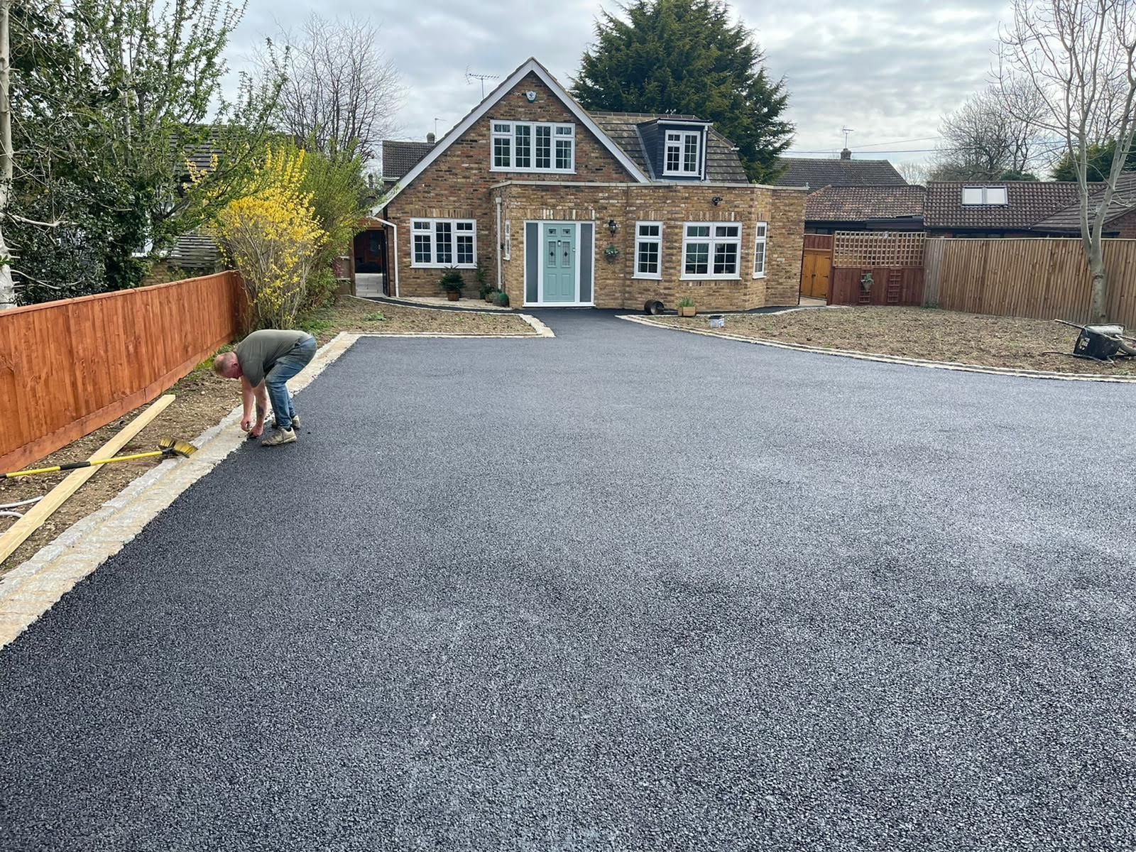 Person working on the edge of a new asphalt driveway in front of a stone house.