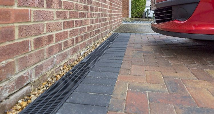 Brick driveway with drain near red car and brick wall.