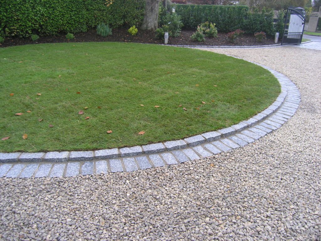 Circular lawn with stone edging, gravel driveway, and hedge in the background.