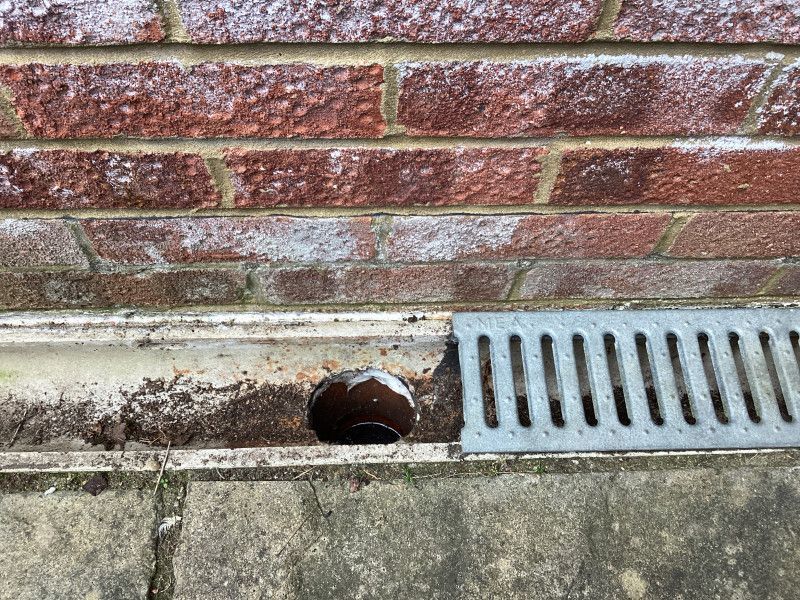 Brick wall above a drainage channel with debris and a metal grate.