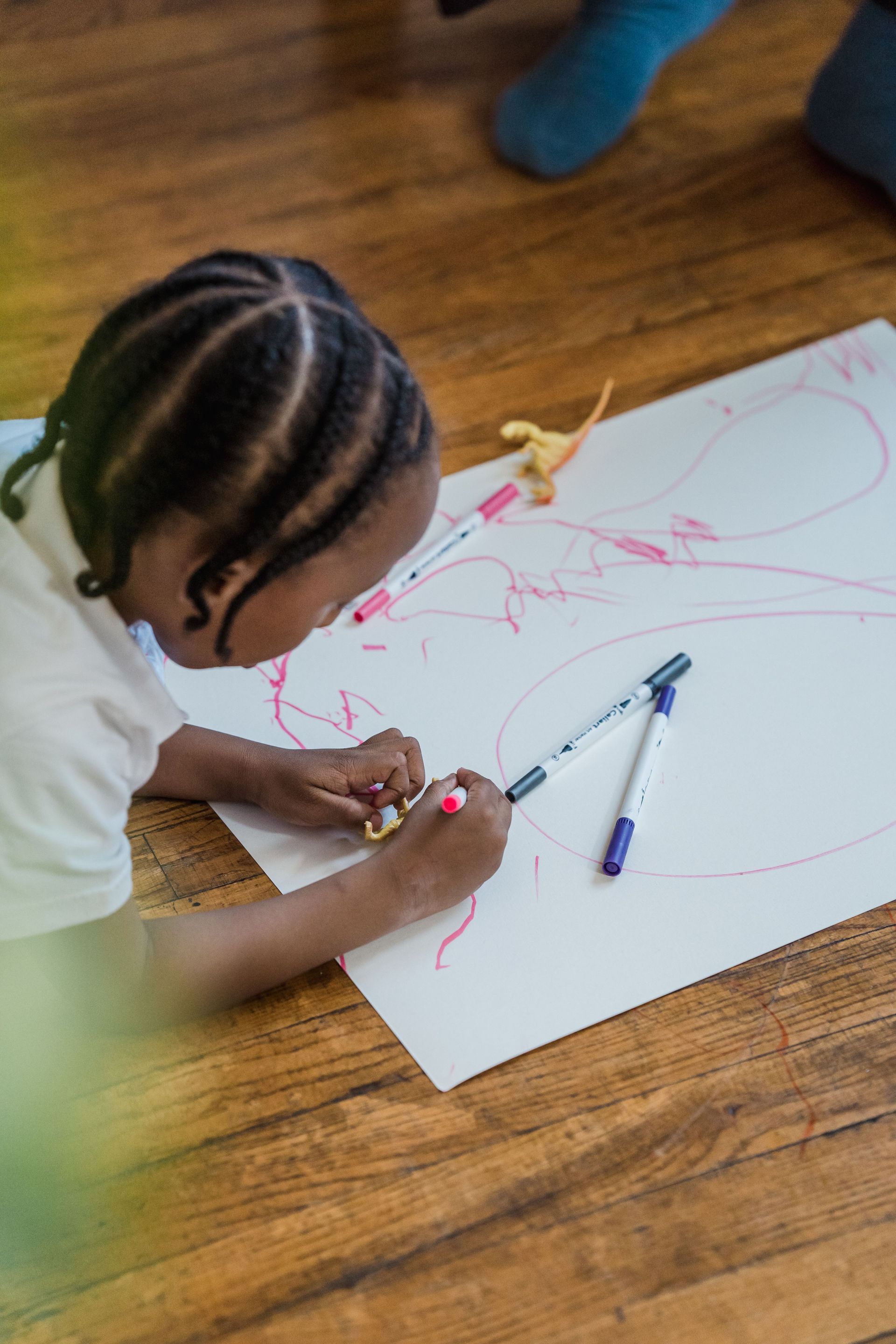 Child drawing with pink crayon on a large white sheet on a wooden floor, with markers nearby.