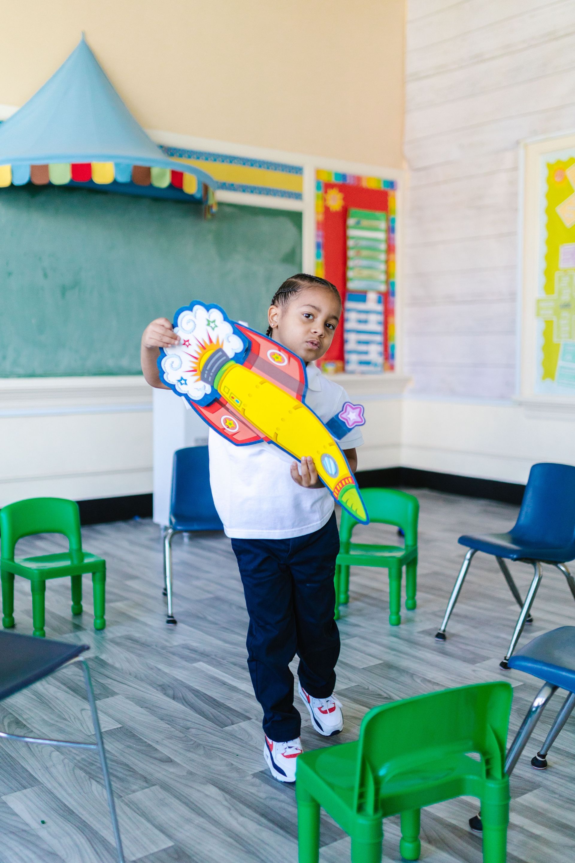 Child holding a colorful toy guitar in a classroom with small chairs and a chalkboard