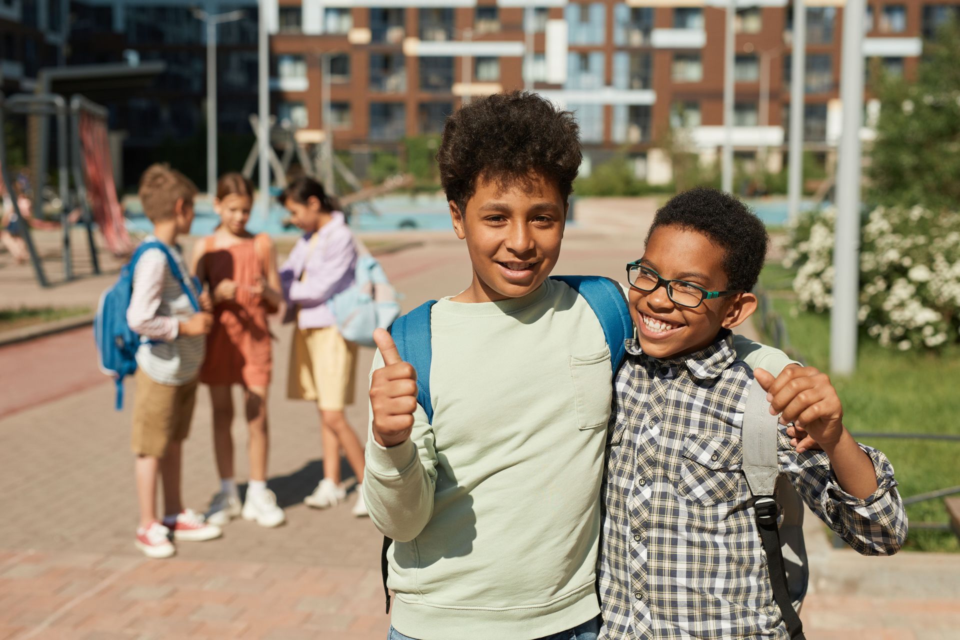 Two smiling children pose with thumbs up in a school courtyard, with classmates blurred in the background.