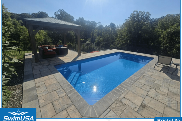 Backyard pool with blue water, stone patio, and shaded lounge area surrounded by trees
