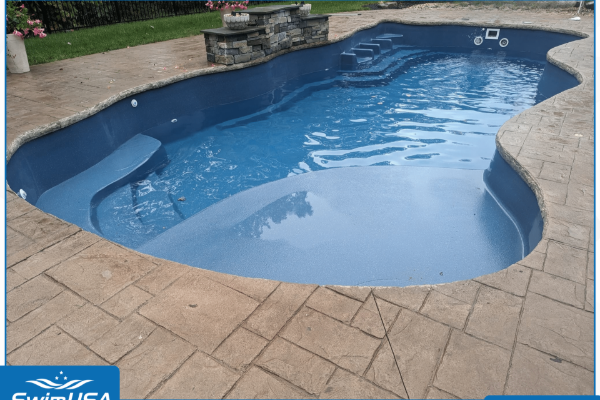 Blue backyard swimming pool with tan stone patio and a small stone waterfall feature