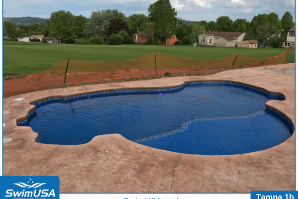 Freeform blue backyard swimming pool with tan deck and green lawn under a cloudy sky