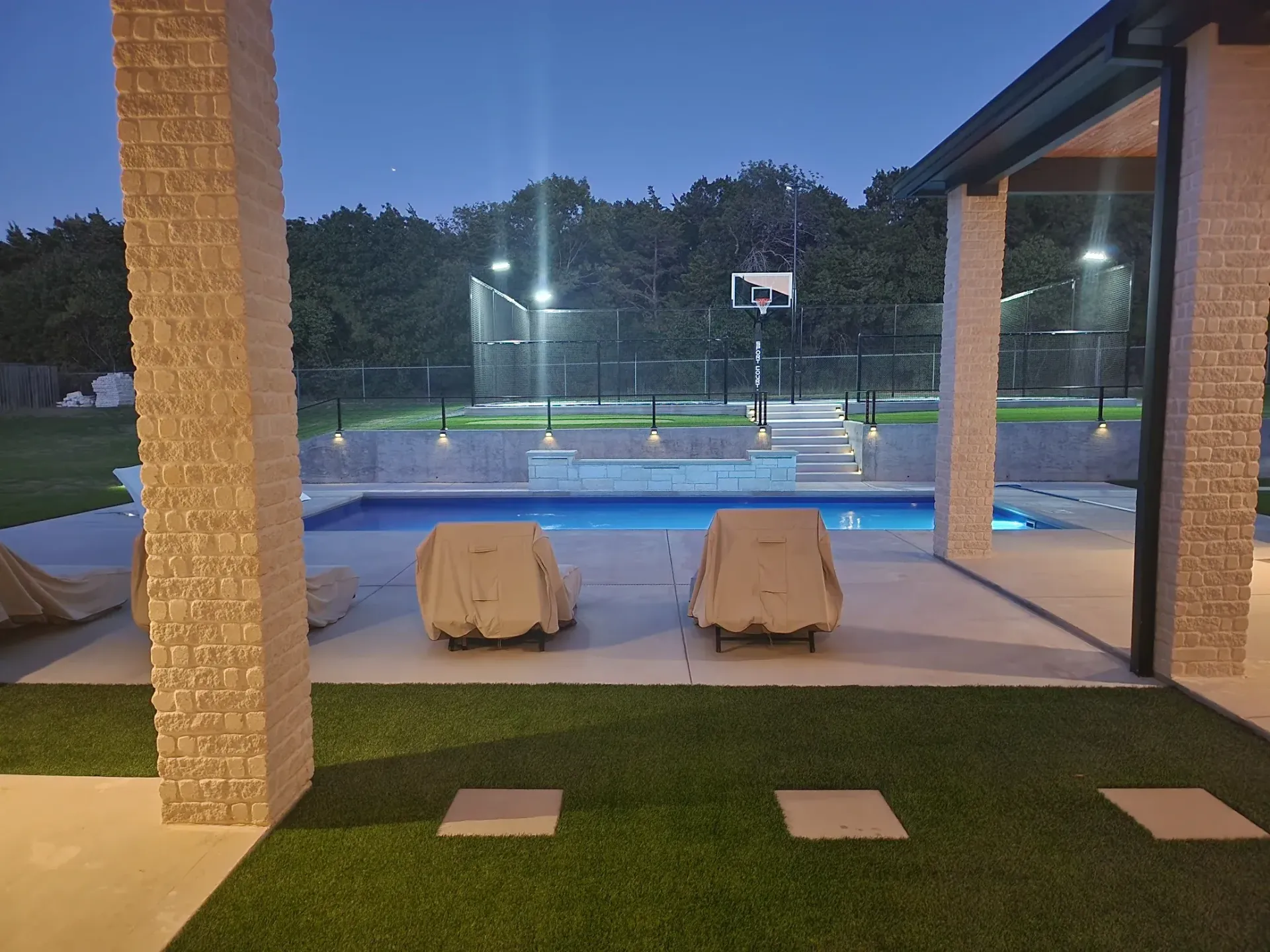 Covered patio overlooking lit tennis court at dusk, with two covered lounge chairs facing the pool.