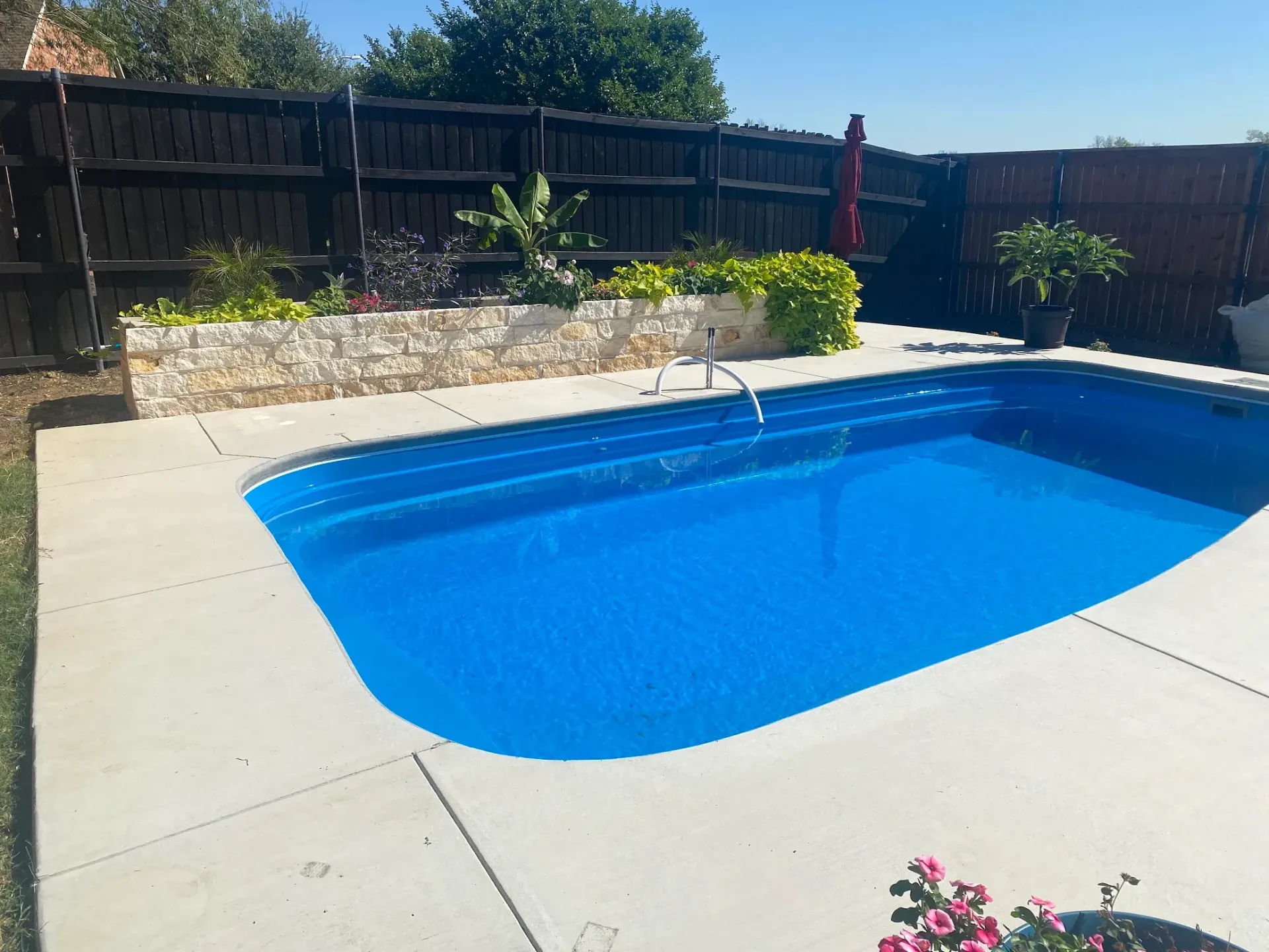 Backyard swimming pool with blue water, stone wall, wooden fence, and patio in bright sunlight
