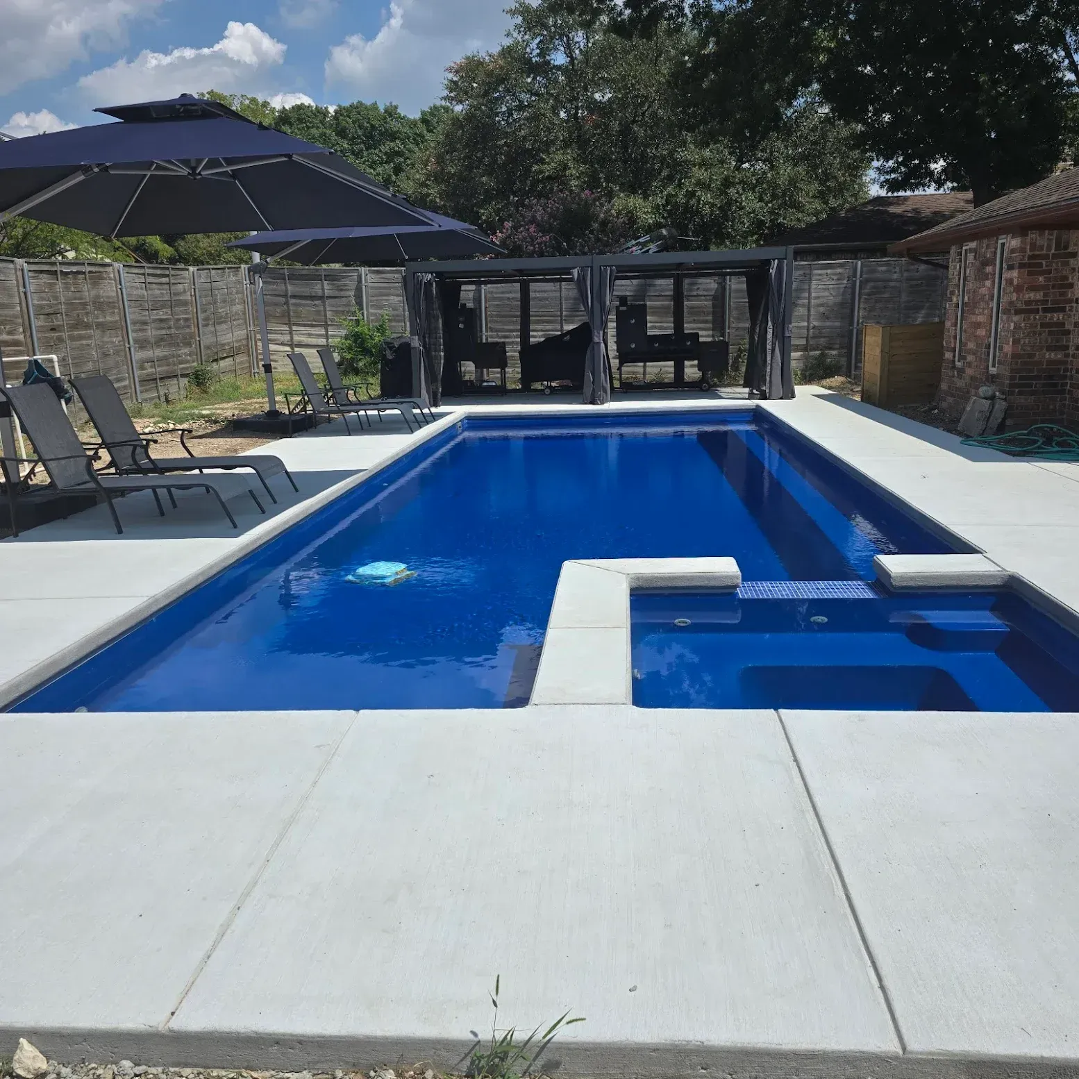 Backyard swimming pool with blue water, white deck, lounge chairs, and umbrellas under a sunny sky