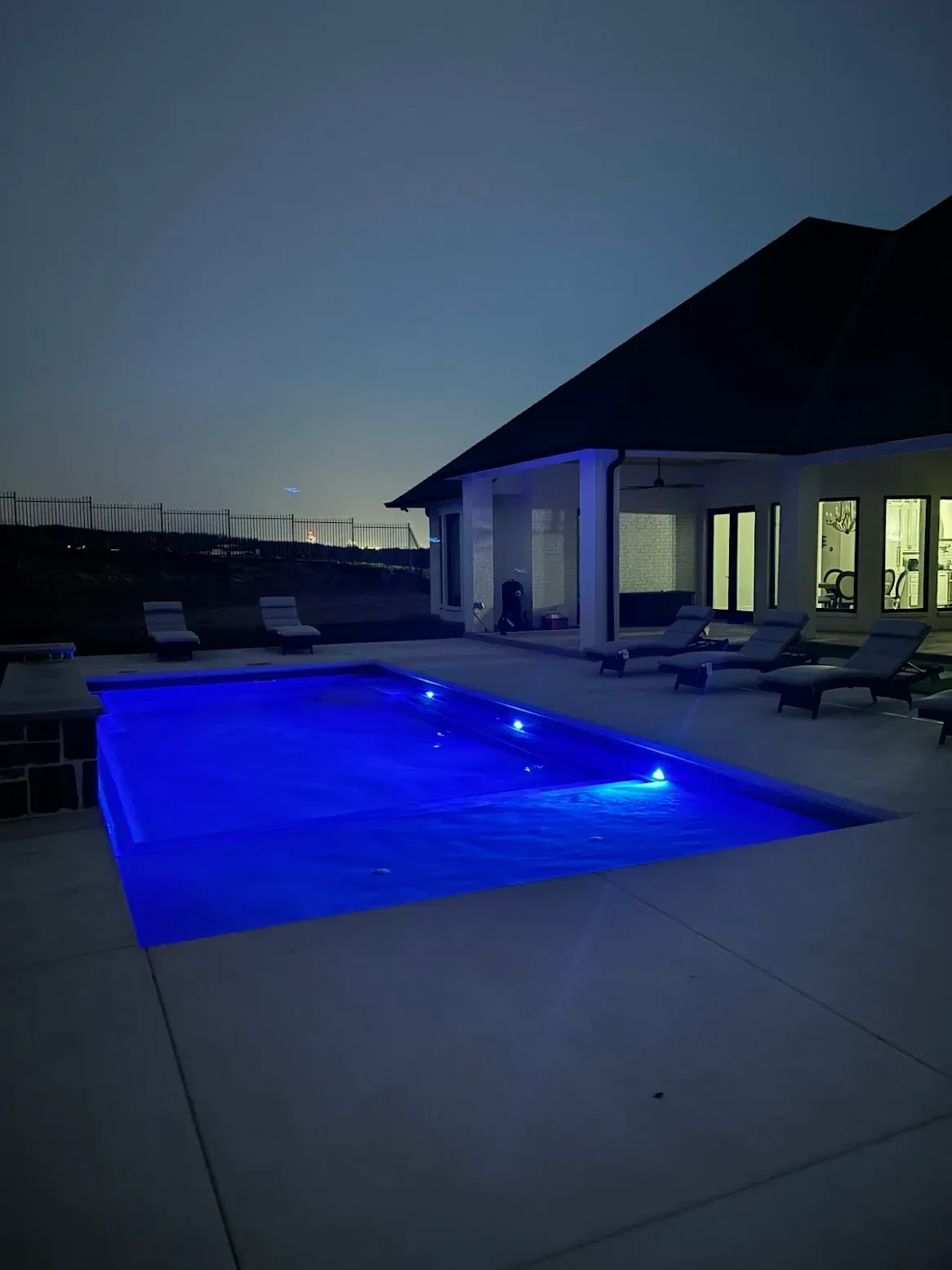 Blue-lit swimming pool beside a modern house at dusk, with lounge chairs on the patio.