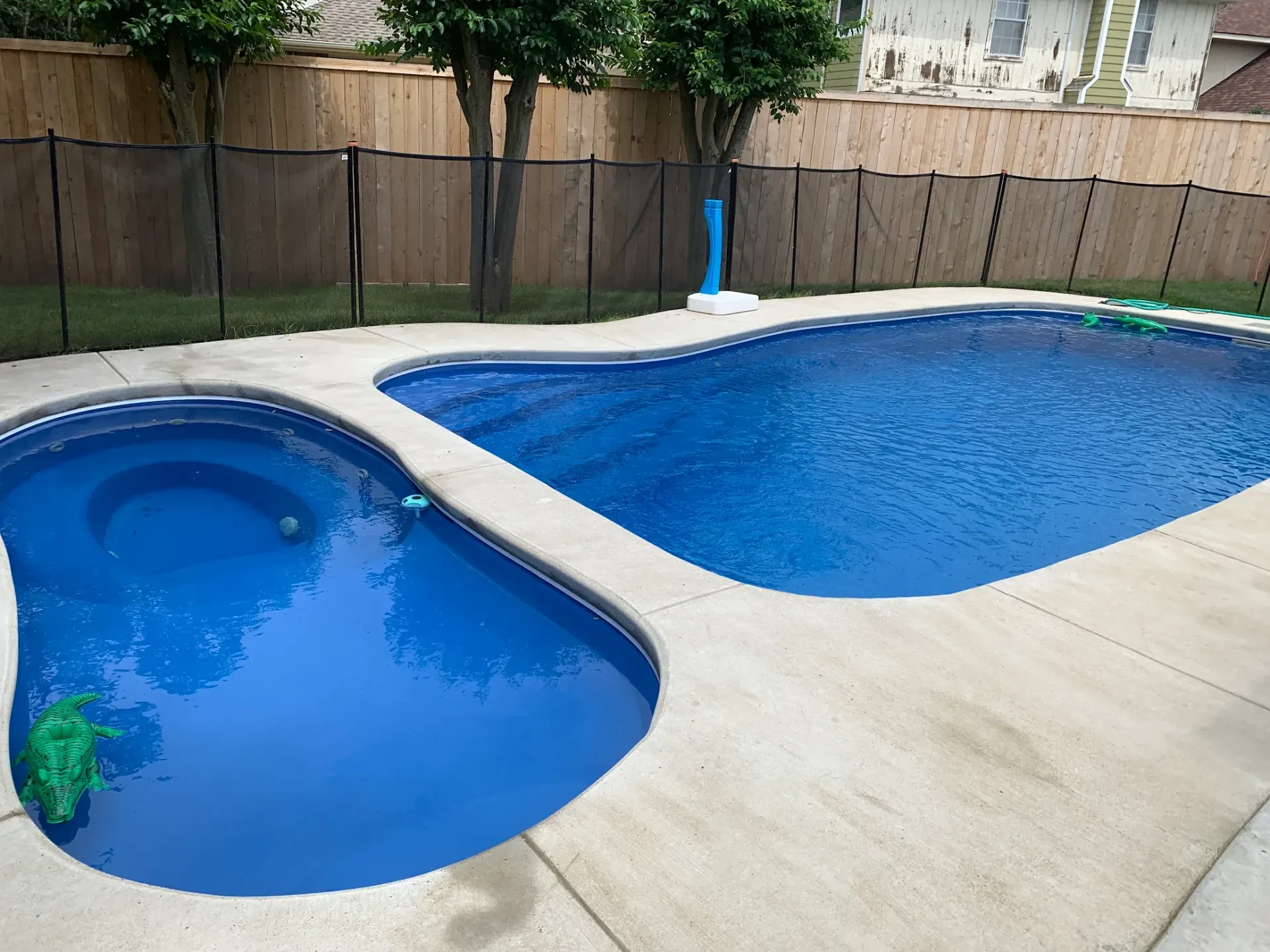 Backyard swimming pool with a small shallow section and a larger blue pool beside a patio and wooden fence