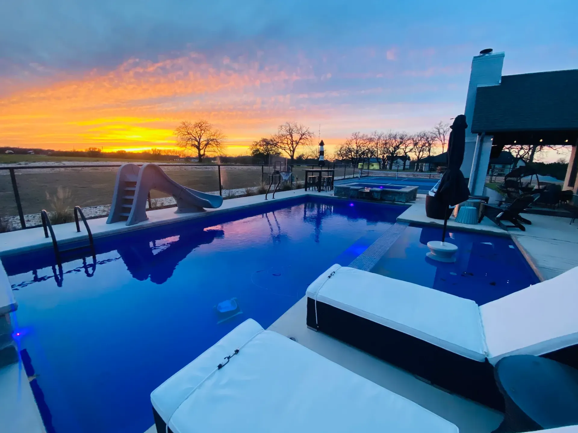 Backyard pool at sunset with blue water, lounge chairs, and orange-purple sky over the horizon