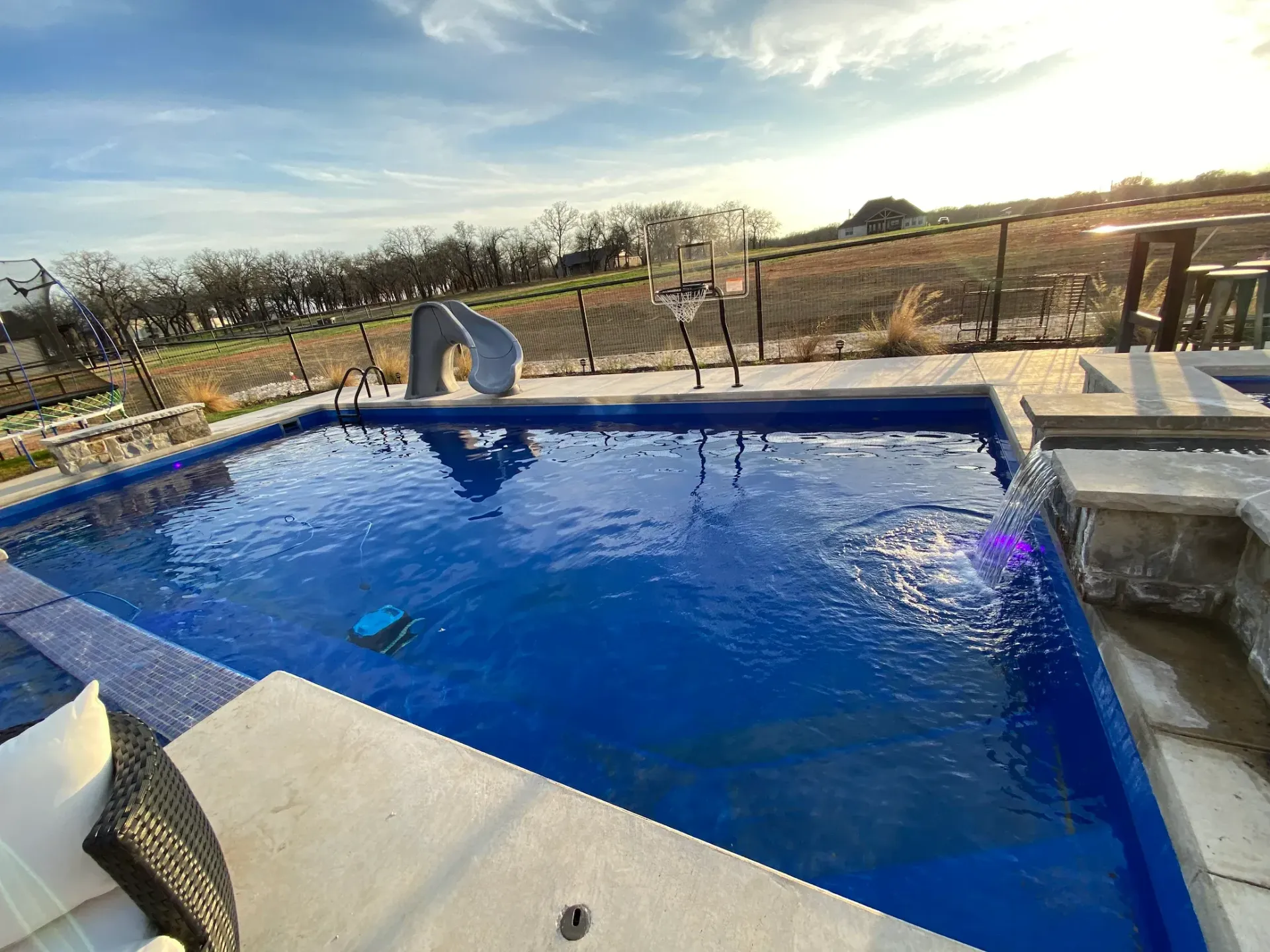 Backyard swimming pool with blue water, stone deck, and hillside view at sunset