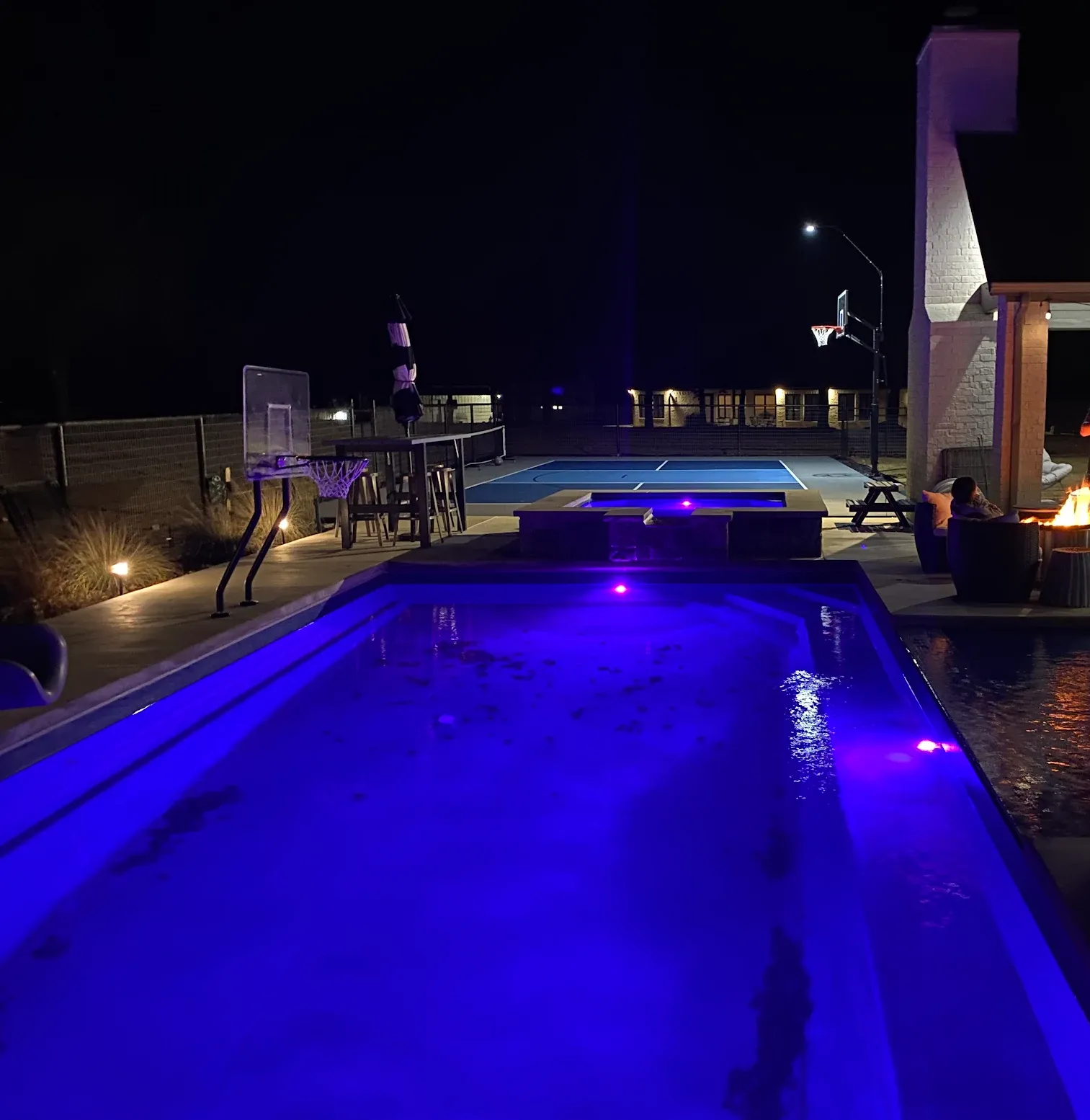 Nighttime backyard with a glowing blue pool, patio furniture, and a lit fire pit