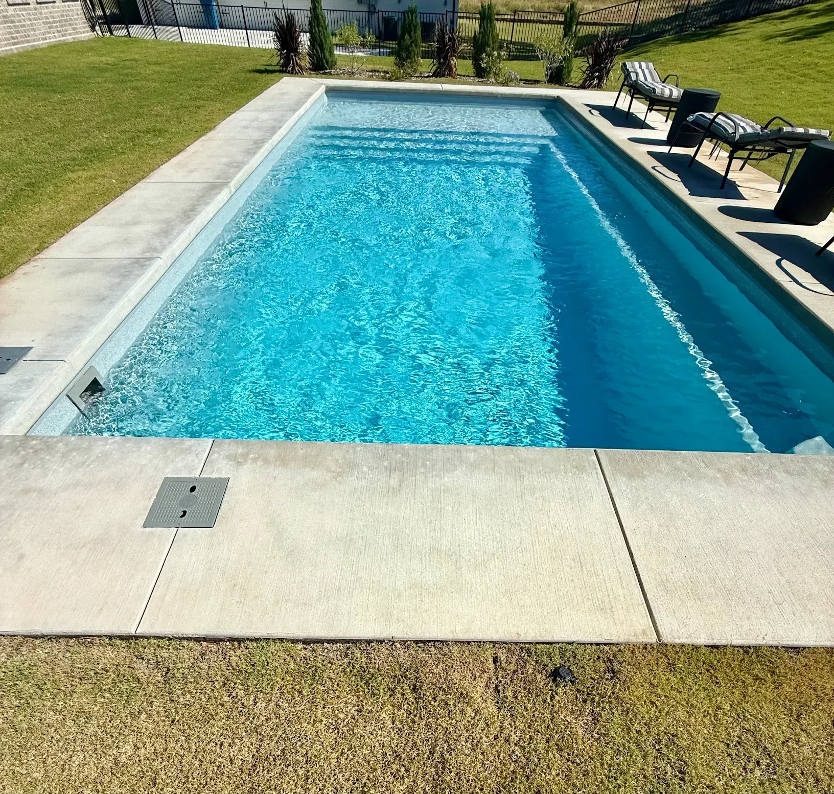 Rectangular backyard swimming pool with blue water, concrete deck, and lounge chairs beside a grassy lawn.