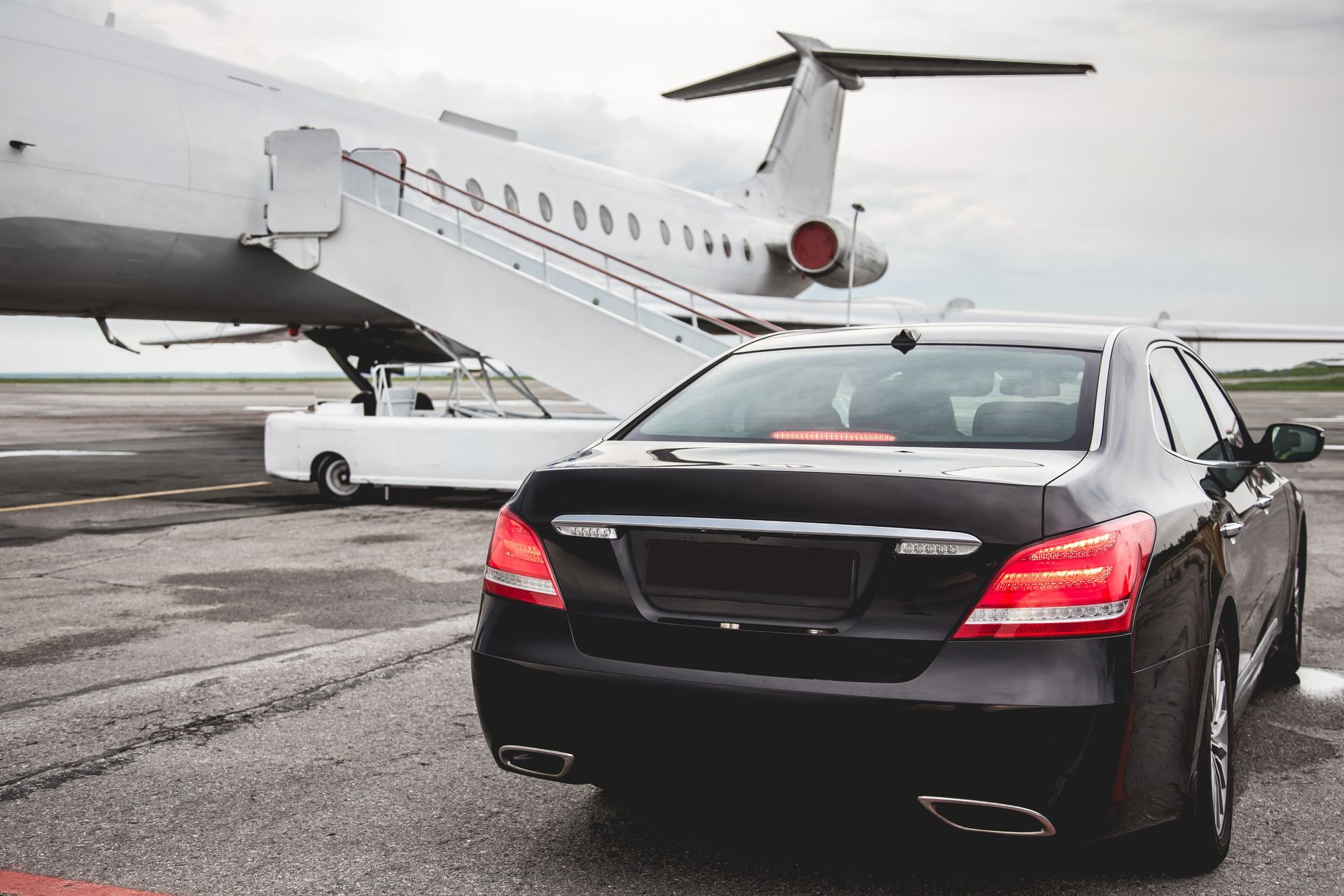 Black sedan parked on a tarmac with a private jet and convertible in the background.