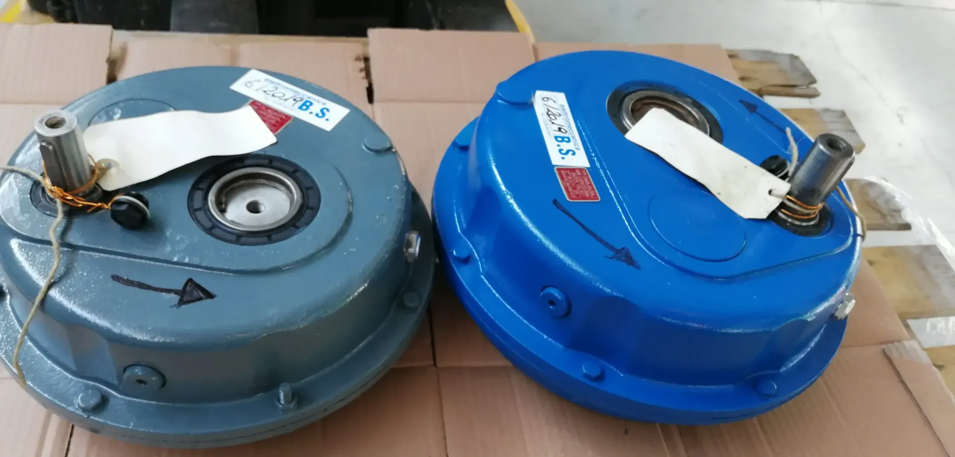 Due componenti industriali, uno grigio e uno blu, con etichette di carta, su una superficie di cartone.
