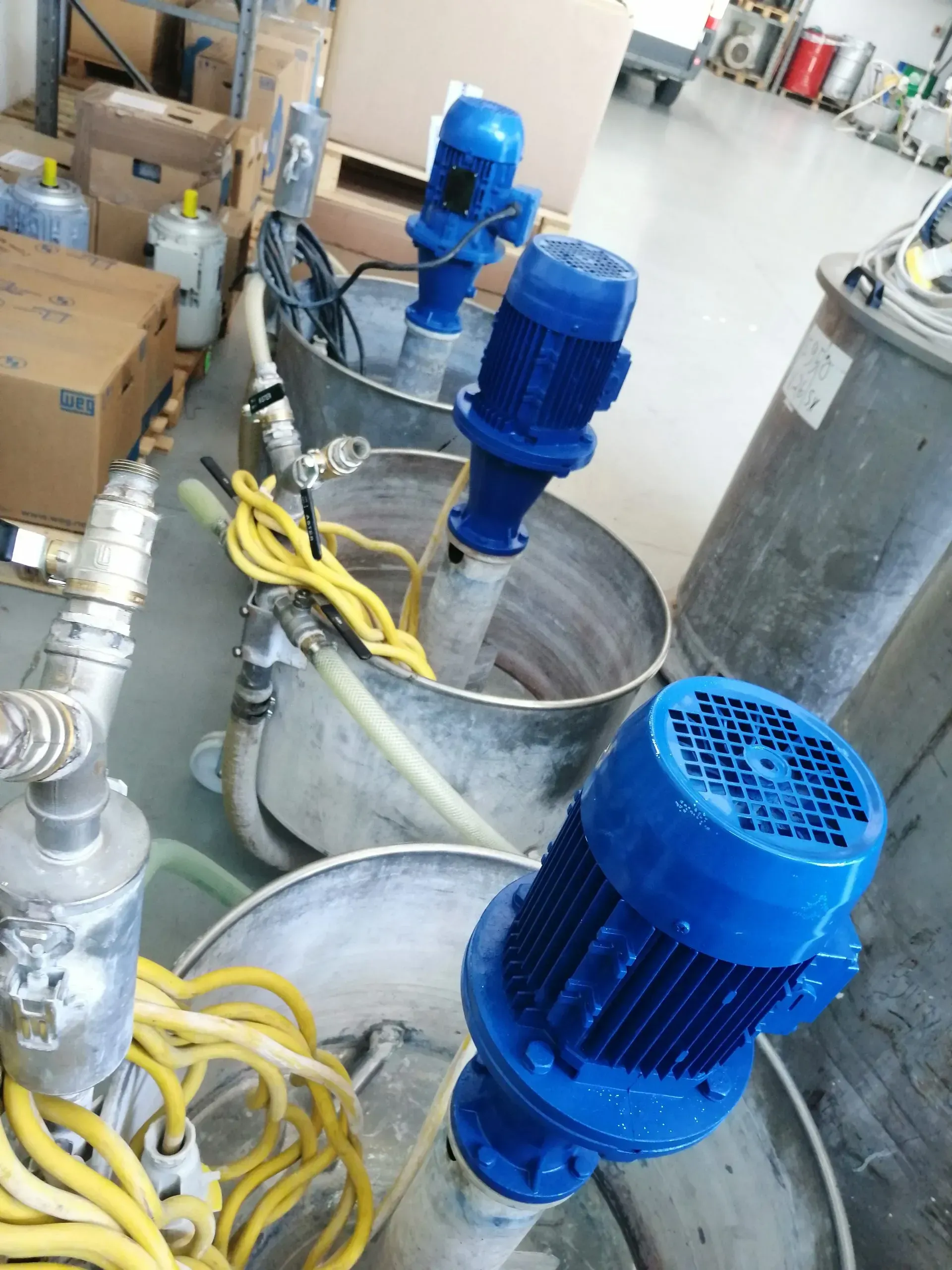 Serbatoi di miscelazione industriali con motori blu, cavi gialli e tubi metallici in un magazzino.