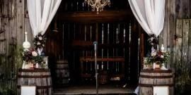 Rustic wedding arch with white drapes, flowers, and wooden barrels in front of a dark backdrop