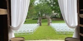 White chairs arranged on a lawn with an outdoor aisle leading to a stone archway, viewed from a tent opening