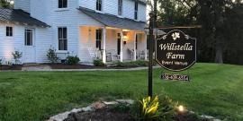 White farmhouse with porch and sign reading “Willstella Farm,” beside a green lawn at dusk