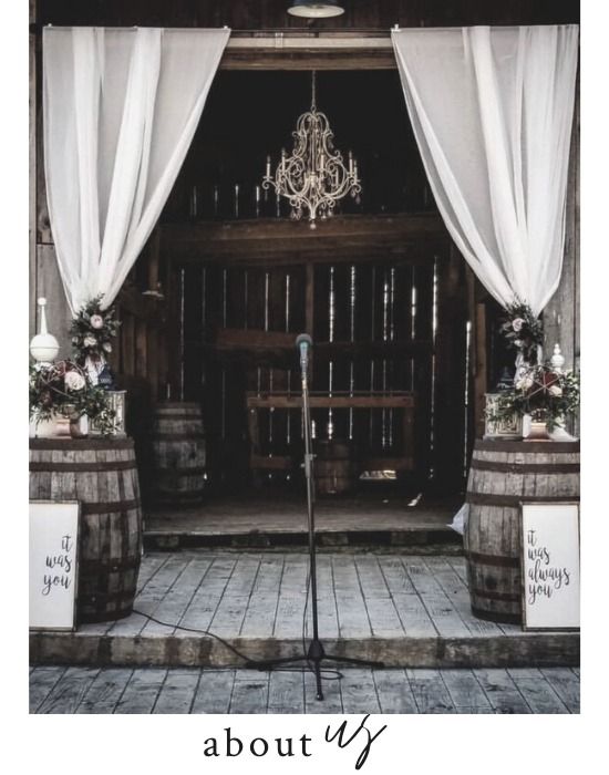 Rustic wedding altar with white drapes, chandelier, and floral arrangements in a barn entrance.