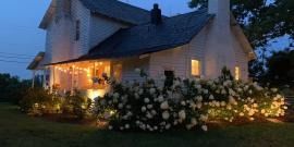 Lit white house at dusk with warm porch lights and blooming flowers in the yard