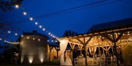 Twilight outdoor wedding reception with string lights, a lit gazebo, and guests under a covered pavilion
