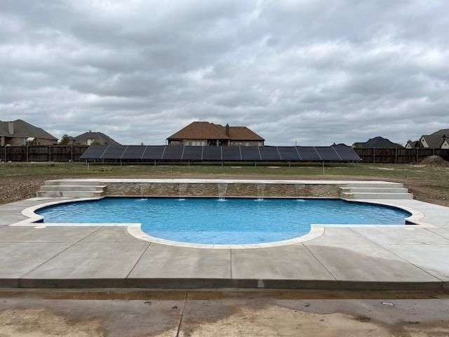 Pool with blue water and waterfall feature. Solar panels line the fence in the background under a cloudy sky.
