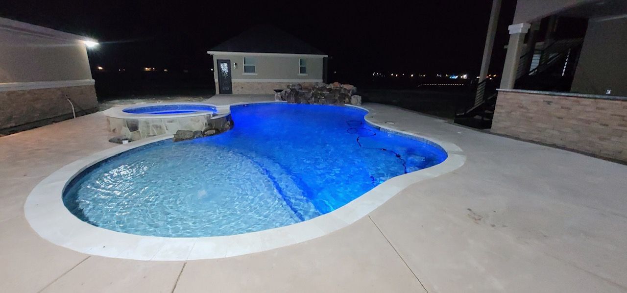 Night view of a pool with blue lights, a hot tub, and a small building in the background.