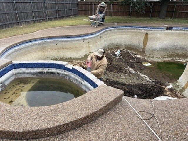 Two people cleaning a drained, dirty swimming pool. One works in the pool, the other with a wheelbarrow. Brown and blue tile.