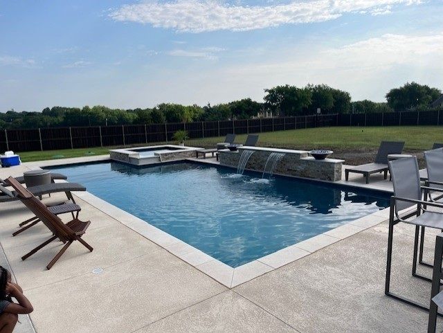 A rectangular swimming pool with a connected jacuzzi, a waterfall feature, and surrounding concrete patio.