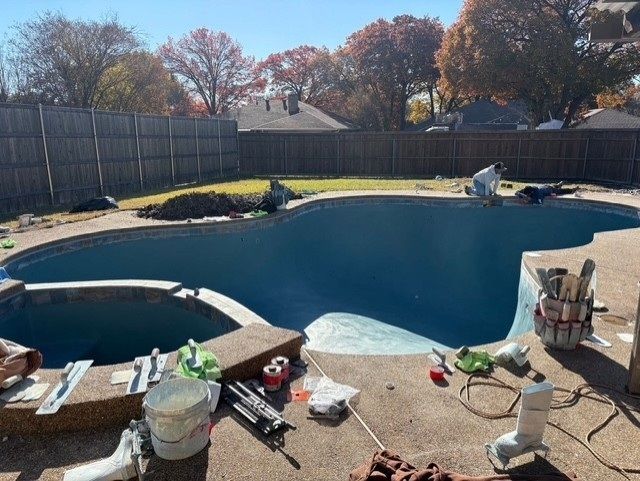 Pool under construction; blue interior, surrounded by patio. Tools and materials scattered around. Fence and trees in background.