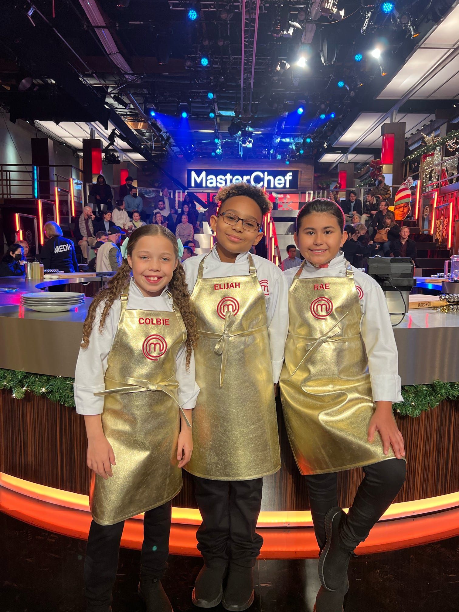 Three people in gold aprons pose on a MasterChef set with the logo lit in the background.