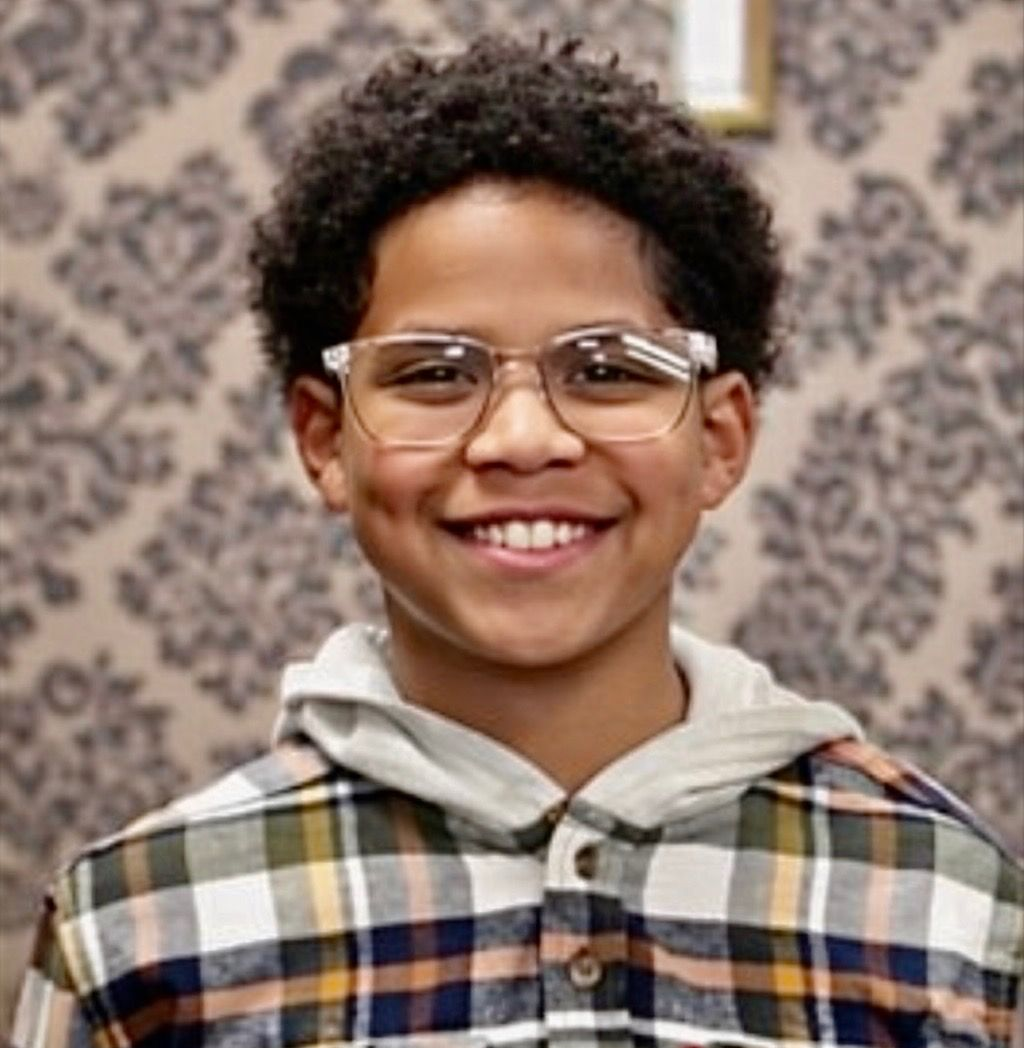 Kaleb wearing glasses and a plaid shirt against a patterned wallpaper background