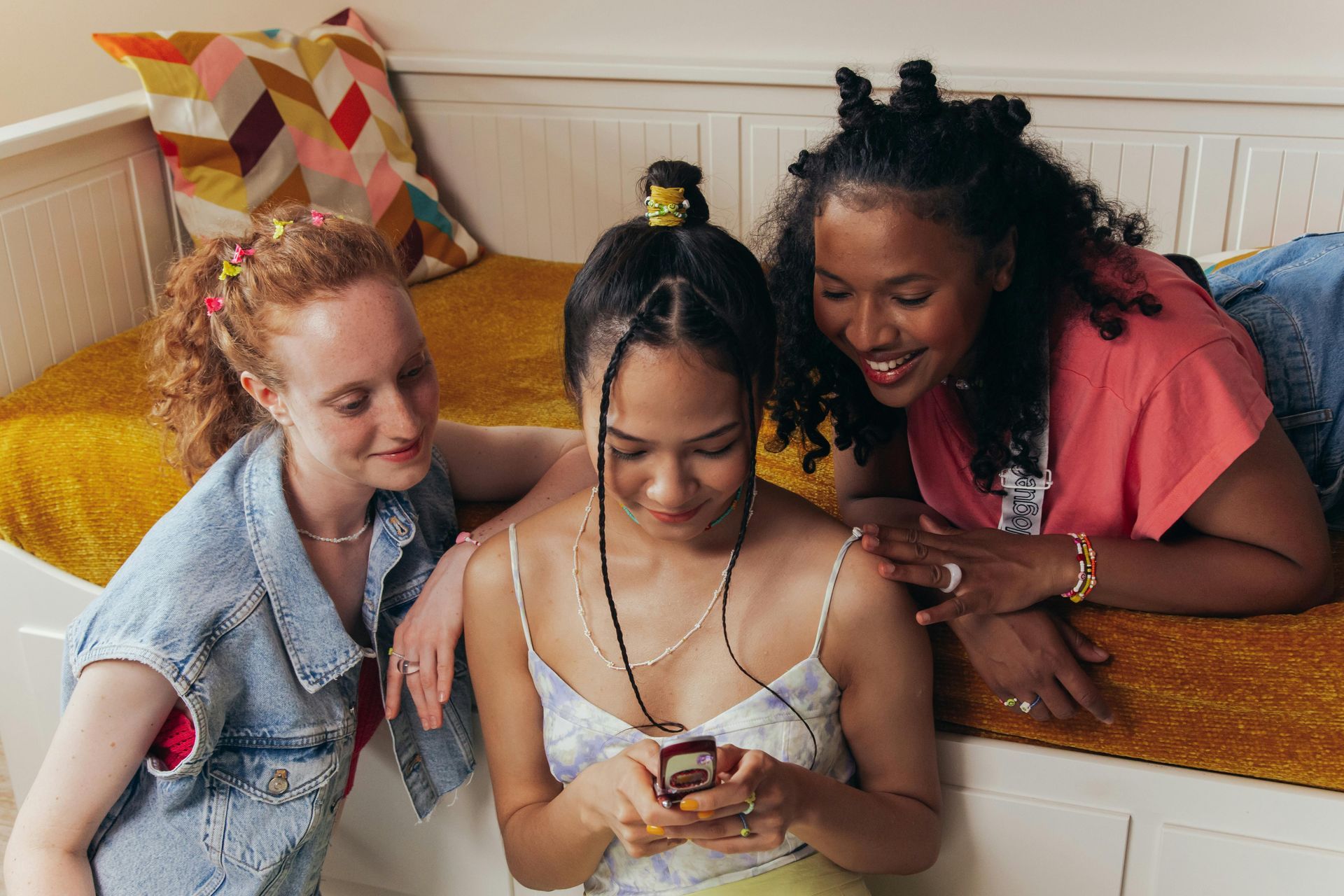 Three friends smiling over a phone on a couch with colorful pillows
