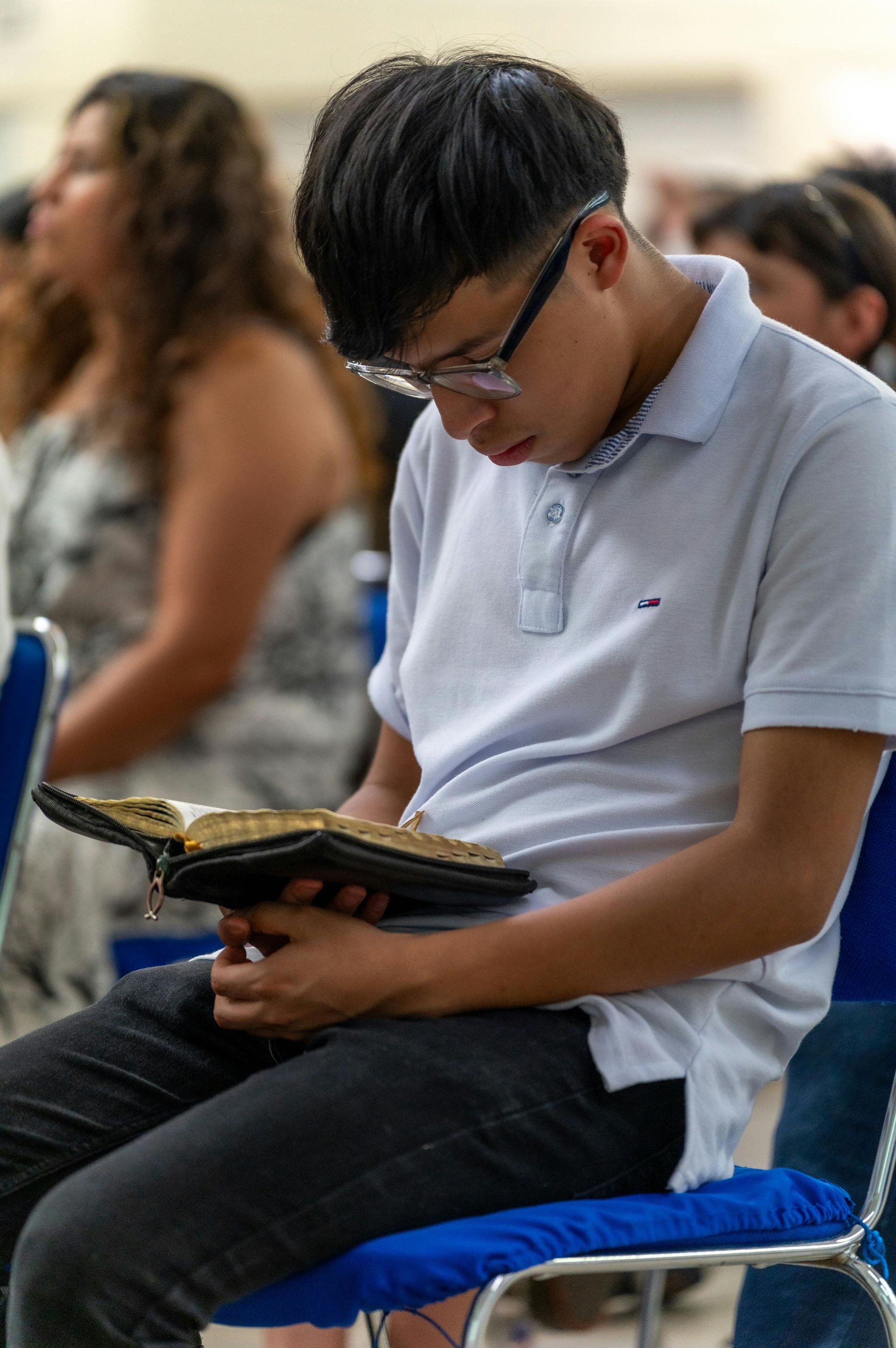 Person in a white shirt sits on a blue chair, looking down at an open book in a classroom setting.