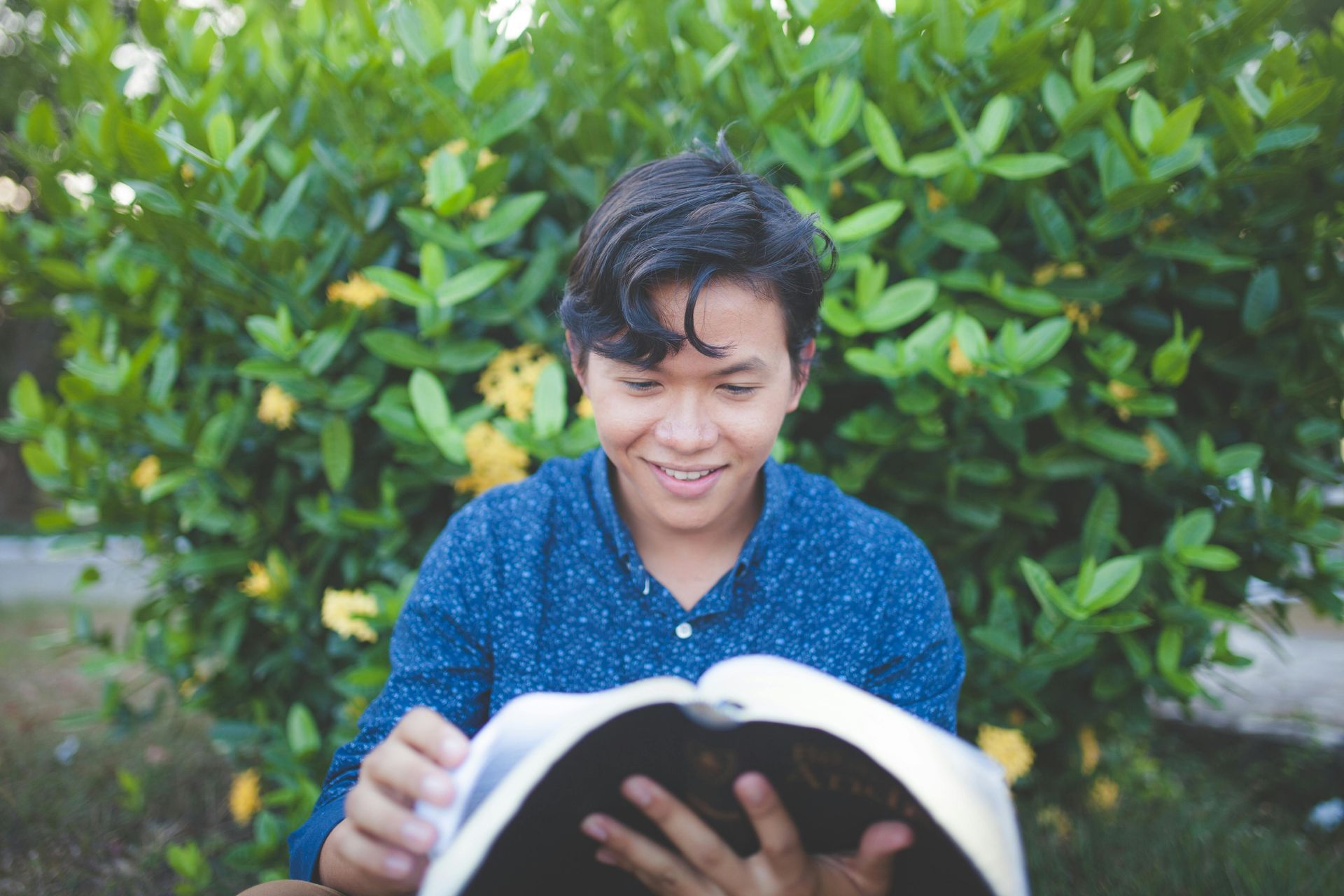 Boy reading a book outdoors in front of green foliage, smiling in a blue shirt