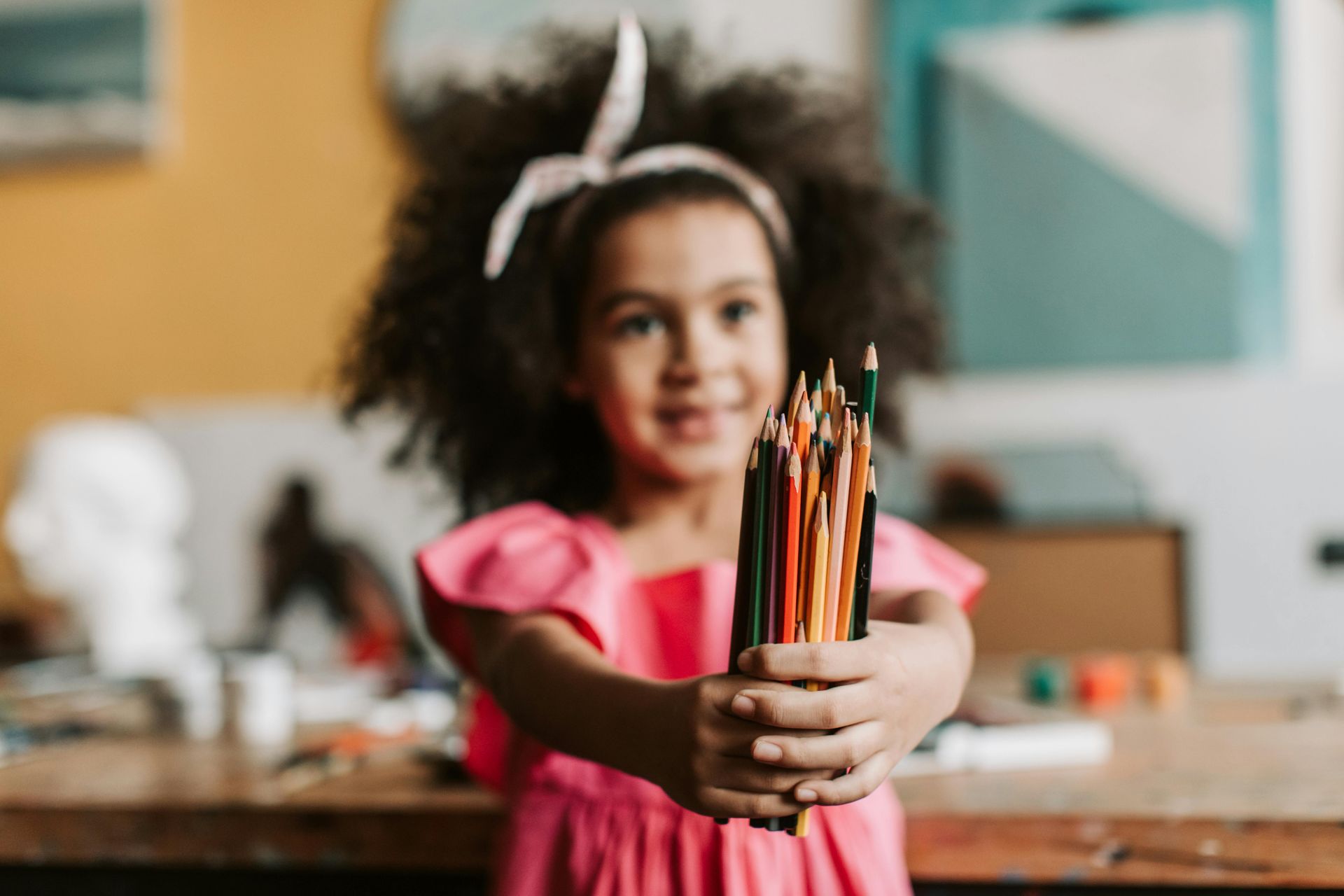 Child in a pink dress holding a bundle of colored pencils, smiling in a classroom