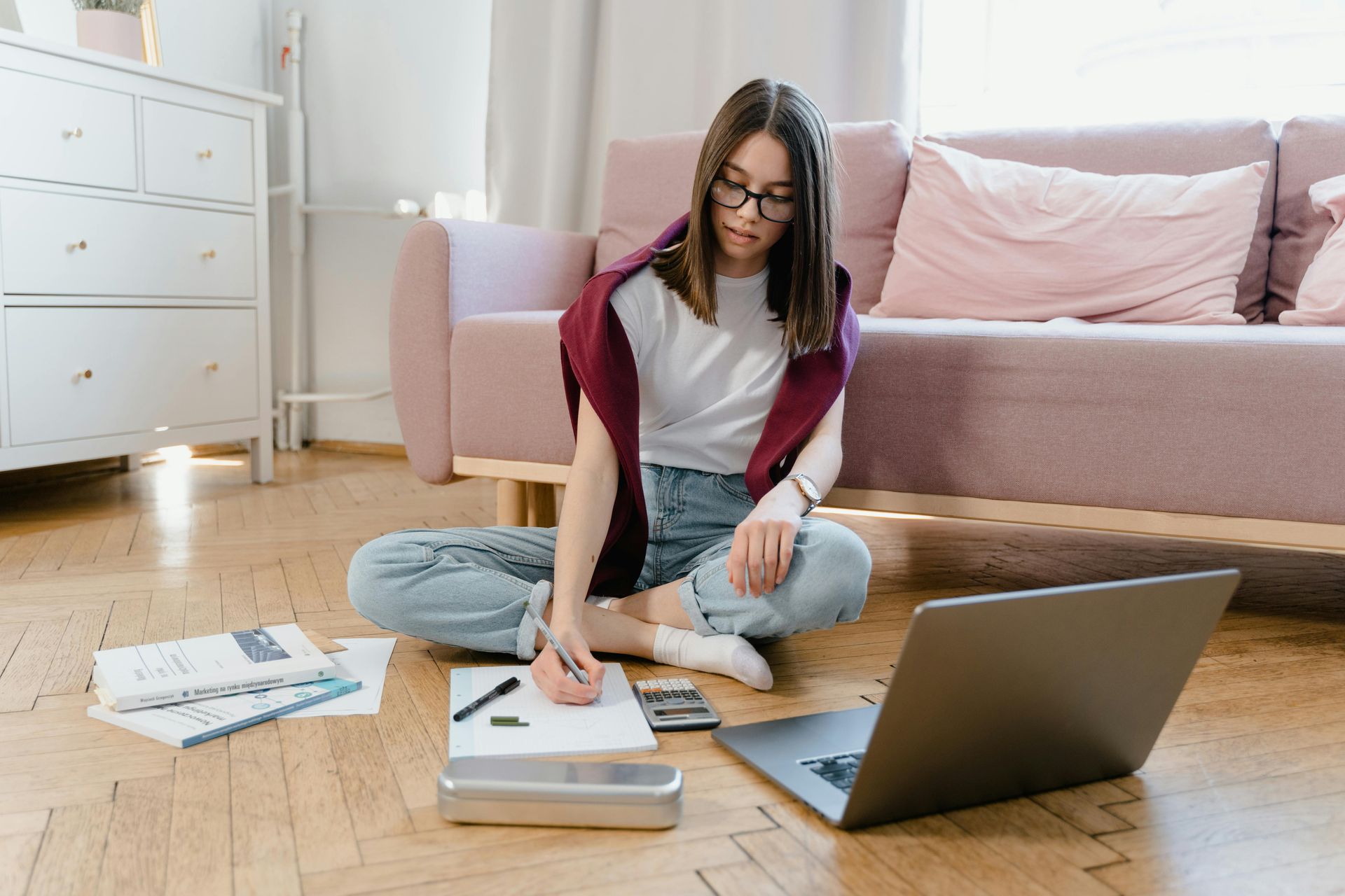 teenager sitting on the floor, writing on papers beside a laptop in a bright living room