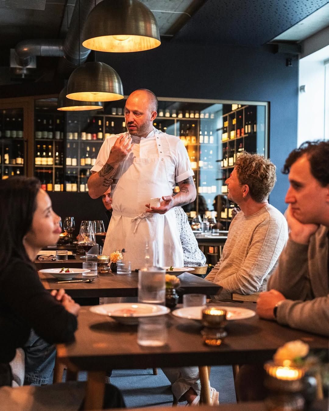 Chef presenting a dish to diners in a dimly lit restaurant.