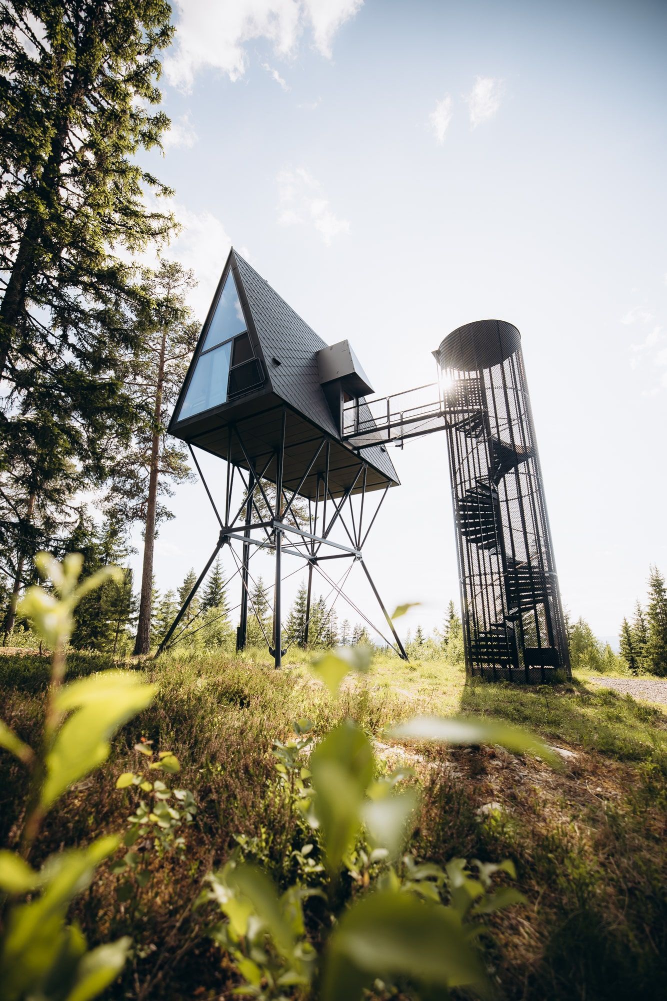 Elevated geometric cabin connected to a spiral tower by a walkway, set in a forest, bright sunlight.