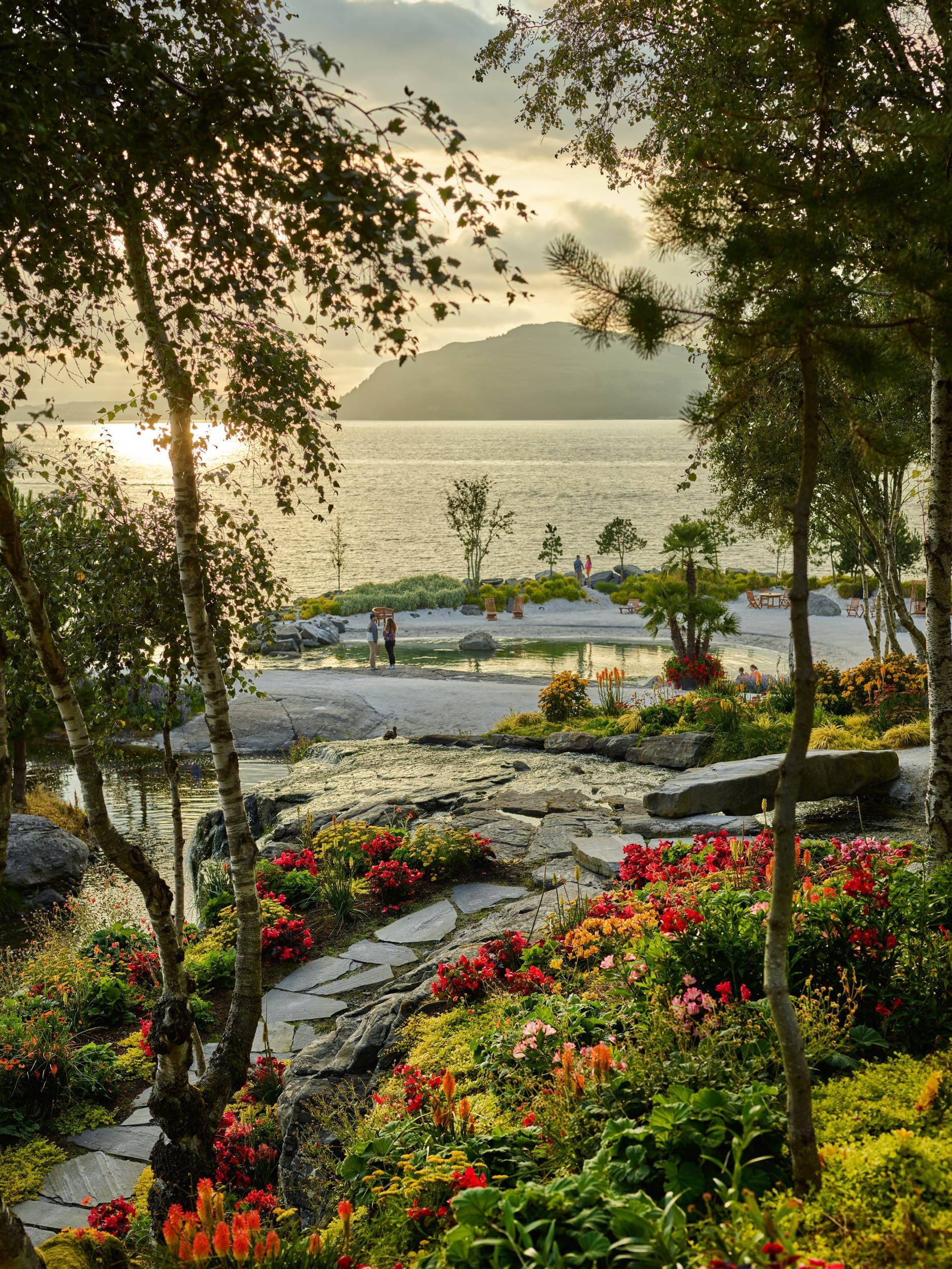 Lush garden with vibrant flowers and stone path overlooking a lake and distant mountain at sunset.
