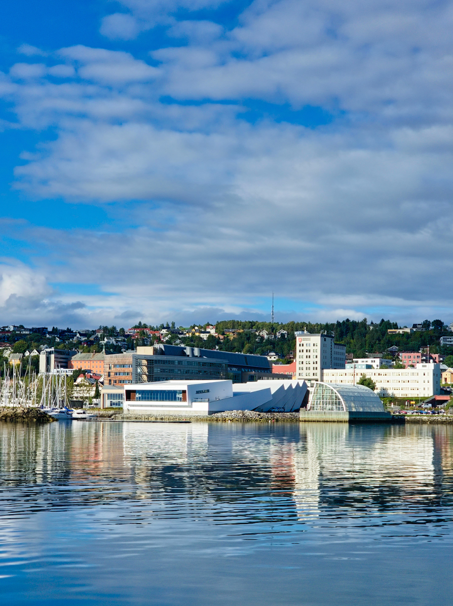 Cityscape with buildings reflecting in the calm water under a blue sky with clouds.