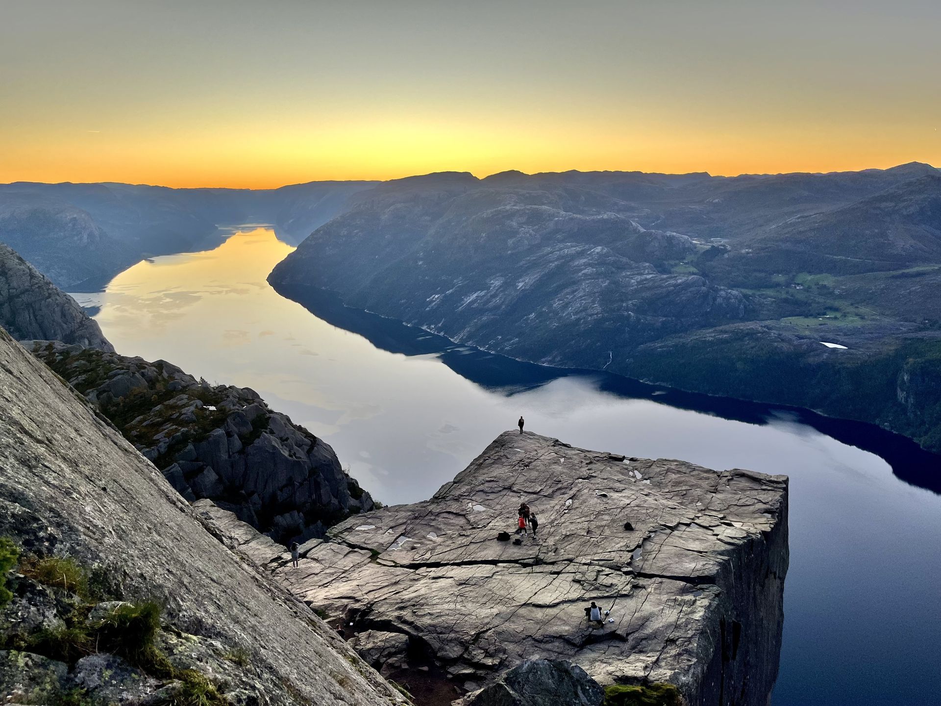 Cliff edge overlooking a fjord. Two figures stand on the rock, with a sunset sky in the background.