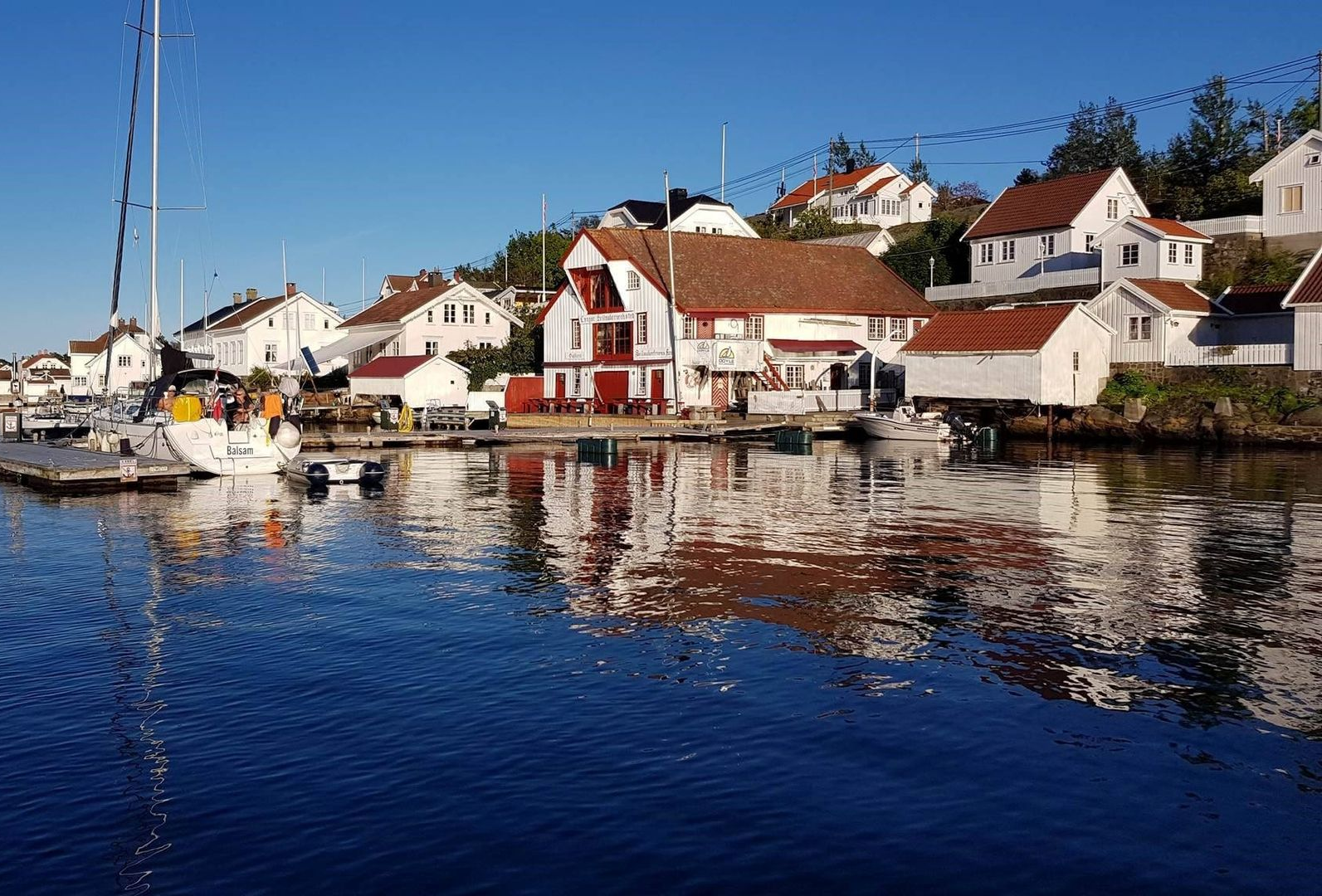 Houses and docks line the water; reflections in the blue water. Bright buildings with red roofs. Sunny day.