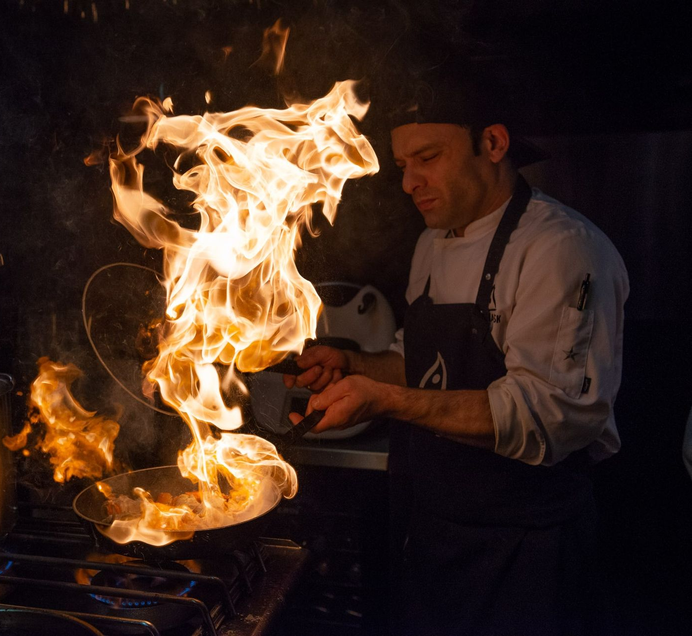 Chef flambéing food in a pan, flames rising high above a gas stove in a restaurant kitchen.
