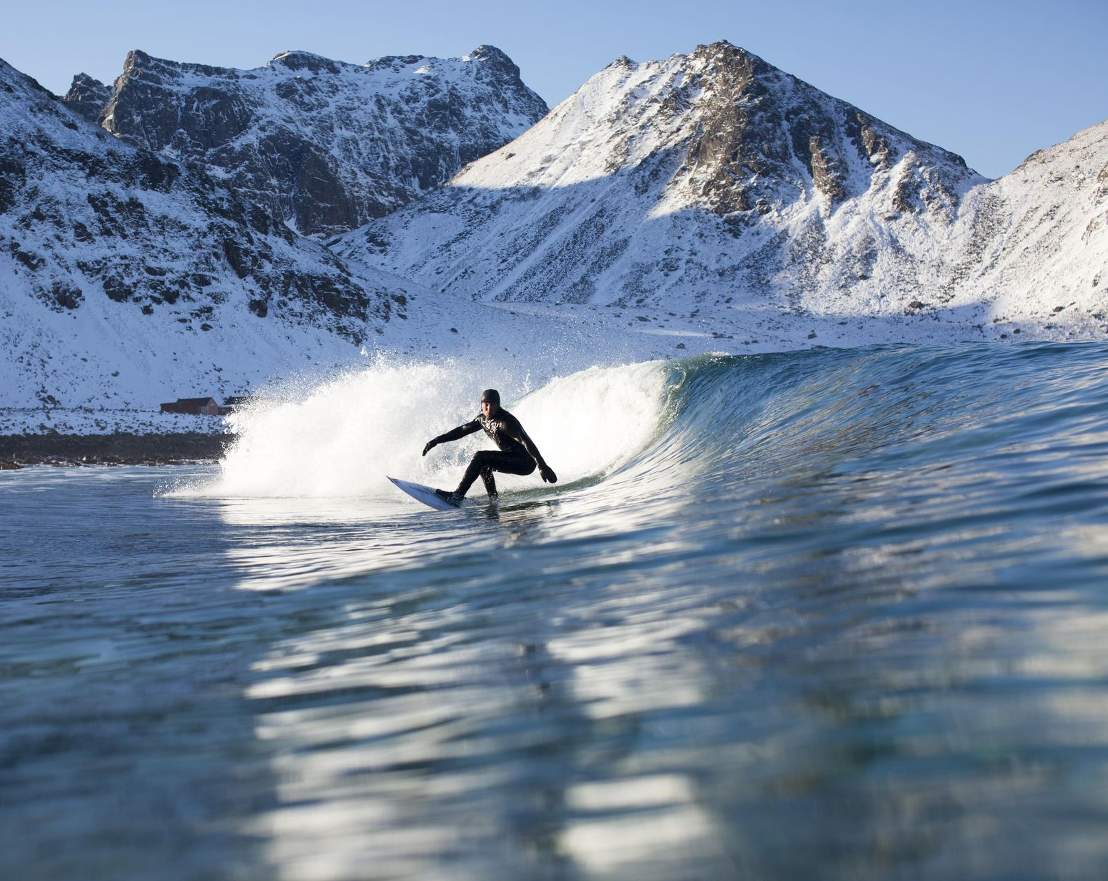 Surfer rides a wave in front of snow-covered mountains. Water is blue; the surfer wears a black wetsuit.