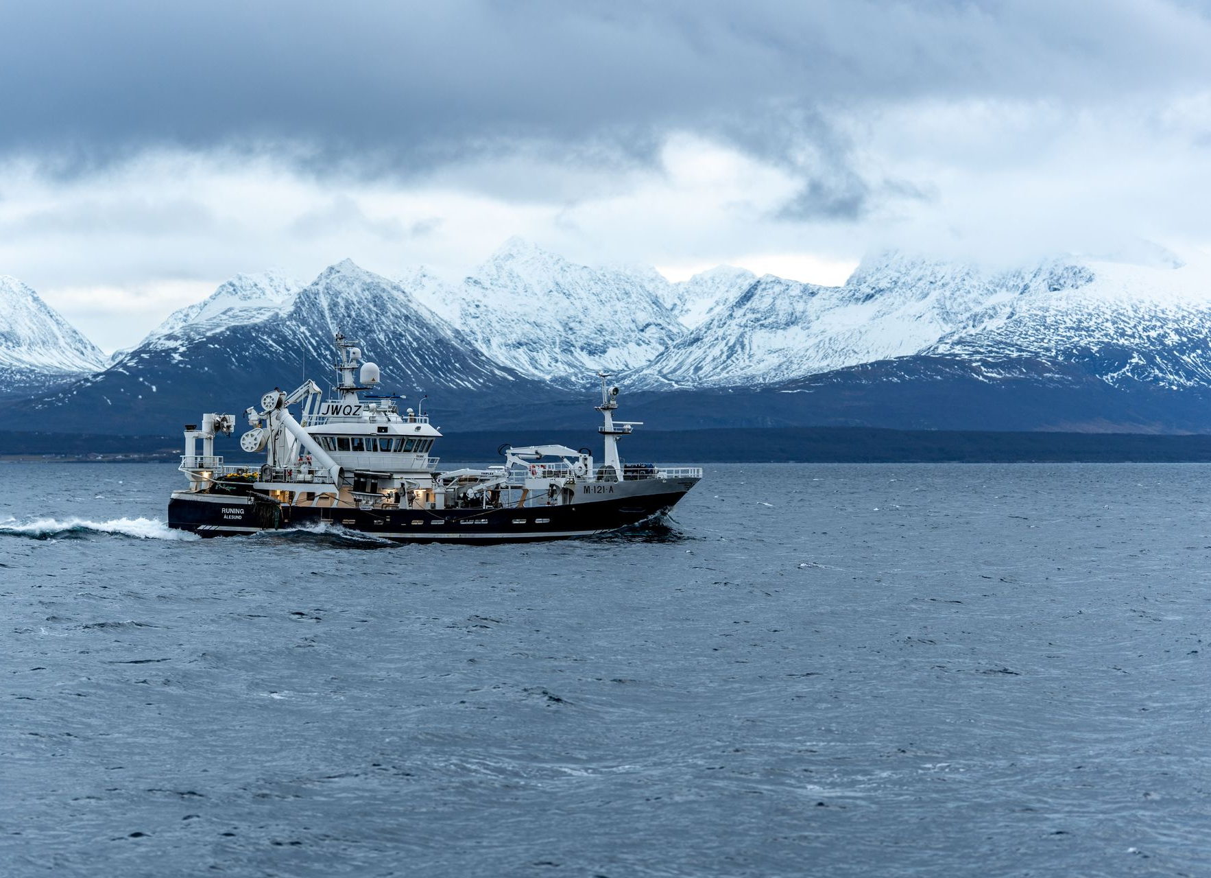 Boat sailing on water with snowy mountains in the background under a cloudy sky.